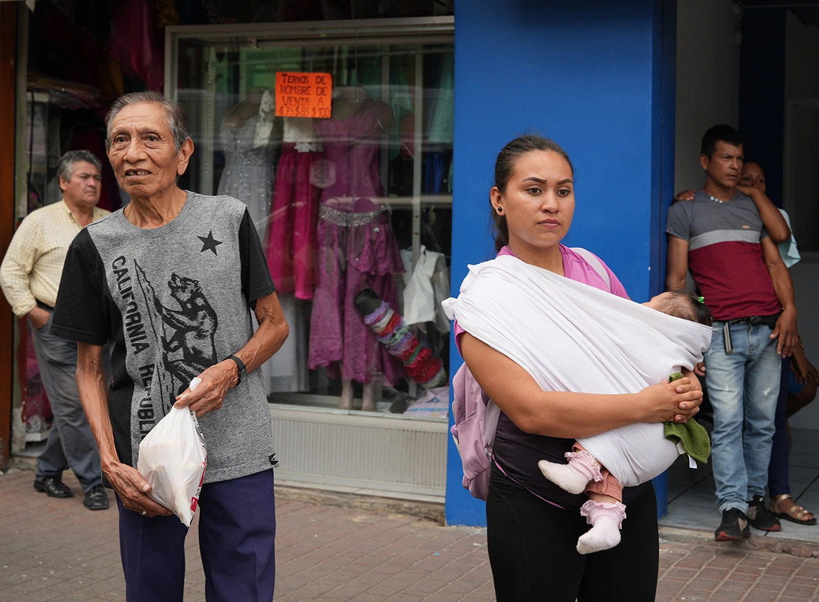 personas en la acera una mujer cargando una bebe un señor con una funda